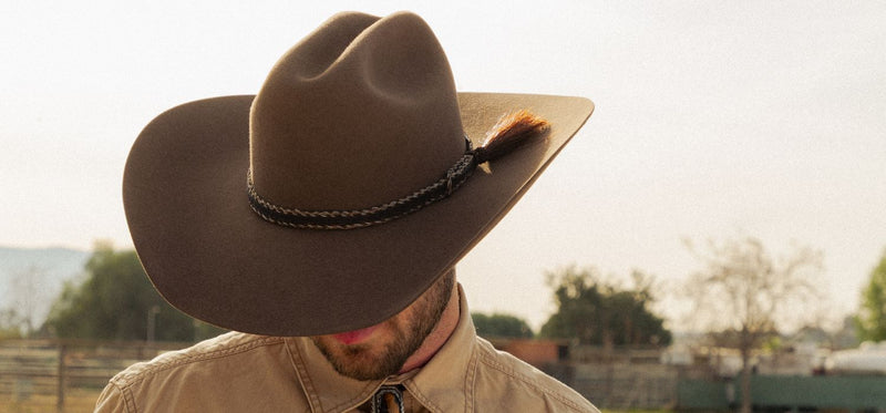 Man wearing a brown cowboy hat with a braided band and feather accent, with the brim shading his face.