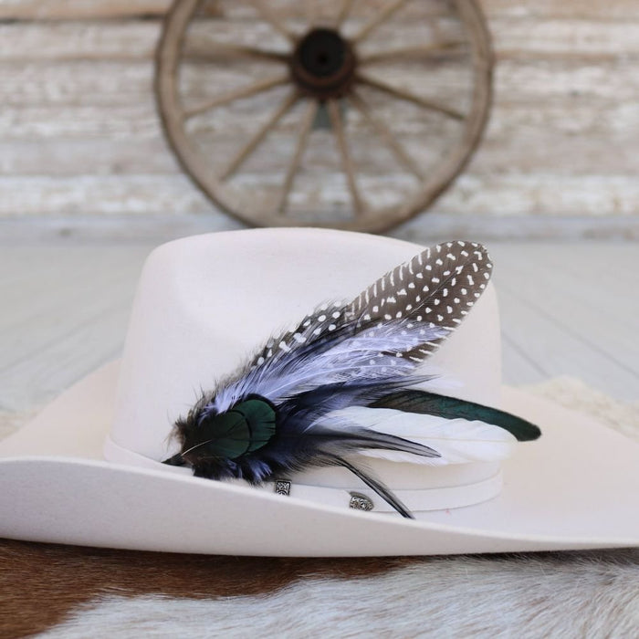 A silver sage feather hat accent tucked into the band of a white hat, displayed against a rustic background.