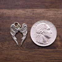 Sterling silver angel wings charm with heart cutout, shown beside a coin